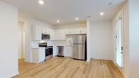 This two-floor space presents a clean, contemporary design characterized by white walls, recessed lighting, and light-toned hardwood-style flooring that flows consistently throughout. The main level features an open-plan layout where the kitchen connects seamlessly to the living area. The kitchen is well-equipped with white shaker-style cabinetry, light countertops, and a full suite of stainless steel appliances, including a refrigerator, electric range, over-the-range microwave, and dishwasher. A glass-paneled entry door and adjacent window allow natural light to brighten the living area, which offers generous open space. A staircase with paneled doors leads to the upper level. The upper floor includes a spacious bedroom with direct access to a private bathroom featuring a dual-sink vanity, light countertops, a walk-in glass-enclosed shower, and hardwood-style flooring. A second full bathroom on this level includes a tub-shower combination and a single vanity. A dedicated laundry area houses a full-sized washer and dryer with overhead wire shelving, adding practical utility to the space. The overall aesthetic is bright, minimal, and move-in ready.
