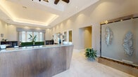 This sophisticated single-floor community space makes a strong first impression with its grand reception area, where a sleek, modern front desk sits beneath soaring ceilings accented by a coffered tray detail with ambient lighting. Polished marble-look tile floors extend through the entry, complemented by an elegant wall-mounted water feature and curated greenery that set a refined tone from the moment of arrival.

The lounge area is a standout, featuring a dramatic floor-to-ceiling stacked brick fireplace wall with a wall-mounted flat-screen TV, flanked by plush leather seating and warm wood-look flooring. Natural light pours in through large windows dressed with layered drapery and wooden blinds. An adjacent gathering space offers a dark-toned dining table beneath two statement lantern pendants, seamlessly flowing into a comfortable seating lounge with built-in countertop workstations along one wall — ideal for both social and productive use.

The kitchen is fully appointed with rich dark cabinetry, a pearlescent tile backsplash, granite countertops, stainless steel appliances, and a casual dining table, making it well-suited for resident events or daily use. Throughout the space, cohesive design elements — including tray ceilings, crown molding, bold artwork, and abundant natural light — contribute to an overall atmosphere of polished, welcoming sophistication.