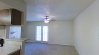 This single-floor space offers a clean and functional layout designed for everyday living. The main living area is finished with soft wall-to-wall carpeting that adds warmth and comfort underfoot. A ceiling fan with an integrated light fixture hangs centrally, providing both air circulation and ambient lighting throughout the room. One of the standout features is the set of French doors, which flood the space with natural light and offer a bright, open feel. The textured popcorn ceiling adds a classic residential character to the interior. Adjacent to the living area, a partial view of what appears to be a countertop or bar surface suggests a connected kitchen or dining area, contributing to the open, flowing nature of the floor plan. The neutral wall tones throughout create a versatile backdrop that complements a variety of interior styles. Overall, the space presents a bright, welcoming atmosphere with practical finishes well-suited for comfortable day-to-day living.