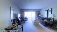 This single-floor residence offers a warm and inviting atmosphere, centered around a generously proportioned living area designed for both relaxation and everyday living. The room is anchored by plush wall-to-wall carpeting that adds comfort underfoot, while neutral-toned walls provide a versatile backdrop for a variety of décor styles. A large sliding glass door with vertical blinds serves as the focal point of the living space, allowing abundant natural light to pour in and offering a connection to the outdoors. The open layout comfortably accommodates a full seating arrangement, including a sofa, accent chair, and an upholstered ottoman that doubles as a coffee table, creating a functional and cohesive gathering space. A dedicated media area features a flat-screen television on a dark wood console, complemented by a classic floor lamp that enhances the ambient lighting. On the opposite side of the room, a dining area with a dark wood table and chairs is visible, providing a seamless transition between living and dining functions. The space is further accented by decorative plants and framed artwork, contributing to a lived-in, homey ambiance throughout.