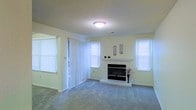 This single-floor space offers a comfortable and well-lit environment designed for relaxed living. The main living area is a spacious, open room centered around a classic fireplace framed by a clean white mantel, serving as a natural focal point for the space. A raised hearth adds a subtle architectural detail, while decorative wall accents above the mantel lend a touch of personality to the room.

Soft, neutral-toned carpeting extends throughout the living area, contributing to a warm and inviting atmosphere underfoot. The walls are finished in a light, airy shade that enhances the sense of openness and brightness. A textured ceiling adds a layer of visual interest overhead.

Natural light pours into the room through multiple windows dressed with crisp white horizontal blinds, creating a fresh and clean aesthetic. A sliding glass door fitted with vertical blinds provides access to an outdoor area, further extending the sense of space and connectivity to the exterior.

Overall, the space maintains a neutral, move-in-ready palette that offers a versatile canvas for a variety of interior styles, balancing practicality with comfortable, everyday livability.