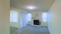 This single-floor space offers a comfortable and well-lit environment designed for relaxed living. The main living area is a spacious, open room centered around a classic fireplace framed by a clean white mantel, serving as a natural focal point for the space. A raised hearth adds a subtle architectural detail, while decorative wall accents above the mantel lend a touch of personality to the room.

Soft, neutral-toned carpeting extends throughout the living area, contributing to a warm and inviting atmosphere underfoot. The walls are finished in a light, airy shade that enhances the sense of openness and brightness. A textured ceiling adds a layer of visual interest overhead.

Natural light pours into the room through multiple windows dressed with crisp white horizontal blinds, creating a fresh and clean aesthetic. A sliding glass door fitted with vertical blinds provides access to an outdoor area, further extending the sense of space and connectivity to the exterior.

Overall, the space maintains a neutral, move-in-ready palette that offers a versatile canvas for a variety of interior styles, balancing practicality with comfortable, everyday livability.