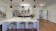 This single-floor community room showcases a cohesive modern farmhouse aesthetic, defined by white shiplap wall paneling and rich hardwood flooring throughout. The open layout connects multiple functional zones, creating a welcoming and versatile gathering space.

The kitchen area serves as a central hub, anchored by a generously sized waterfall quartz island with an undermount sink and upholstered bar stools. Dark cabinetry contrasts with the white shiplap backdrop, while open metal shelving displays decorative accents and small plants. Stainless steel appliances, including a French door refrigerator, complete the space. Matte black pendant lights hang above the island, adding a contemporary touch.

Adjacent to the kitchen, a casual lounge area features a large sectional sofa accented with layered throw pillows and flanked by side tables with lamps. Multiple windows fitted with white blinds allow generous natural light while offering views of an exterior deck. A decorative wall clock adds a farmhouse-inspired focal point.

A secondary seating nook includes a wall-mounted flat-screen television above a dark wood console, accompanied by a plush wingback chair. Additional seating near an interior wall provides a quieter retreat. Recessed lighting and a ceiling fan ensure comfort throughout. A bulletin board near the entrance adds a functional, community-oriented detail.
