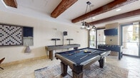 Located at 2121 S Hiawassee Rd in Orlando, FL, this single-floor community game room offers a well-appointed recreational space designed for social engagement and leisure. The architecture draws on Mediterranean-inspired elements, most notably the rich, dark exposed wood ceiling beams that run throughout and contrast warmly against the crisp white walls and ceilings. Underfoot, travertine-style stone tile flooring in a classic pattern adds texture and elegance to the space.

The game room is generously equipped with a full-size pool table centered beneath a stylish industrial pendant light fixture, a shuffleboard table, a foosball table, and a classic Ms. Pac-Man arcade cabinet — offering entertainment for a wide range of interests. A large wall-mounted board game panel and a leather seating area with eclectic décor, including a colorful license plate USA map installation, add personality and character to the space.

Natural light filters in through large glass doors that open to an outdoor corridor featuring arched architectural details, connecting the interior seamlessly to the surrounding grounds. The adjoining lobby hallway is equally inviting, with pendant lantern lighting, potted tropical plants, and wooden accent chairs arranged along the corridor, reinforcing the warm, resort-style ambiance of the overall facility.