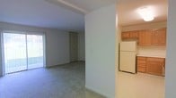 This single-floor space features a straightforward, functional layout that connects the main living area to the kitchen. The living room is finished with wall-to-wall carpet in a neutral gray tone, providing a comfortable underfoot feel throughout the space. A sliding glass door fitted with vertical blinds allows natural light to filter in while offering access to the outdoors. The walls are painted in a clean, light neutral color, contributing to a bright and airy atmosphere.

A partial wall divides the living area from the kitchen, creating a semi-open flow between the two spaces. The kitchen features vinyl flooring, warm-toned wood cabinetry mounted above the counter, and a white full-size refrigerator. The layout appears compact yet practical for everyday use.

Structural beam detailing is visible along the ceiling of the living area, adding a subtle architectural element to the otherwise simple design. A door visible in the background of the living area suggests access to additional rooms or closet storage. Overall, the space presents a clean, move-in-ready environment with a no-frills, practical aesthetic suited for comfortable everyday living.