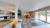 This single-story community clubhouse in Vacaville, California, is designed as a vibrant, multifunctional gathering space that balances comfort and recreation. The interior is defined by a contemporary aesthetic, combining bold color accents with neutral tones throughout. Geometric-patterned carpeting in shades of gray and blue anchors the main open area, while recessed lighting illuminates the space evenly.

The lounge area is furnished with plush orange sofas, a rustic wood coffee table, and colorful accent pillows, creating a warm and inviting atmosphere. A stacked stone accent wall and a wall-mounted fireplace add a tactile, natural element to this seating zone. Adjacent to the lounge, a game area features a foosball table and a wall-mounted flat-screen television set against a bold teal accent wall, with globe pendant lighting overhead.

The kitchenette area offers a white dining table surrounded by green chairs, a refrigerator, sink, and countertop space accented by a dark subway tile backsplash with a decorative open shelf. Sliding glass doors open directly to an outdoor pool area, filling the space with natural light and providing easy access to exterior amenities. Large windows throughout ensure the interior remains bright and connected to the surrounding landscape.