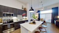 This thoughtfully designed single-floor residence showcases a seamless open-concept layout that connects the kitchen, dining, and living areas into one cohesive living space. Soaring ceilings amplify the sense of openness throughout, while large windows flood the interior with natural light.

The kitchen is a standout feature, outfitted with rich dark cabinetry, crisp white quartz countertops, a tile backsplash, and a full suite of stainless steel appliances including a French door refrigerator, range, microwave, and dishwasher. A generously sized central island with seating provides both additional prep space and a casual dining option, accented by stylish pendant lighting overhead.

The living area continues the bright, airy aesthetic with soaring walls that accommodate dramatic artwork displays and large windows dressed with floor-to-ceiling curtains, creating an inviting atmosphere.

The bedroom offers a comfortable retreat, featuring plush carpeting, a ceiling fan, and ample natural light from well-sized windows with decorative drapery. The adjoining bathroom mirrors the home's modern design language with dark cabinetry, white quartz countertops, a large vanity mirror, and wood-look flooring. Throughout the residence, consistent use of warm wood-tone flooring, clean white walls, and contemporary fixtures creates a cohesive and polished aesthetic.