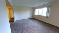 This single-story home offers a clean, classic aesthetic defined by bright white walls and trim throughout. The layout accommodates three bedrooms and one and a half bathrooms, providing a practical and comfortable floor plan suitable for a variety of living arrangements.

The kitchen is well-equipped and features white cabinetry with decorative scalloped detailing along the upper cabinet edges, offering ample storage space. Appliances include an electric range, a white refrigerator, and a dishwasher. Neutral laminate countertops complement the cabinetry, while a window above the sink allows for natural light. Warm hardwood flooring grounds the space and adds character.

The living areas are carpeted in a neutral, multi-toned plush carpet that carries through multiple rooms, contributing to a cohesive and comfortable feel underfoot. The main living room features a large window that brings in natural light and provides outdoor views of the surrounding landscape. An adjacent space, likely a dining area, is accessed through an open archway and is illuminated by a ceiling-mounted light fixture. White wainscoting detail adds a touch of classic charm to this area. The overall ambiance is clean, bright, and move-in ready.