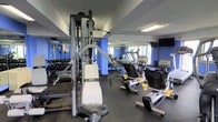 This fitness center offers a comprehensive selection of exercise equipment arranged across a single-level space with a functional, no-frills layout. The room features rubberized flooring suitable for heavy equipment and high-impact workouts. Strength training is well-represented, with a Precor cable machine and a rack of dumbbells lining one mirrored wall, allowing users to monitor their form during exercises. The mirrored walls also help create a sense of additional depth and openness within the space.

On the cardio side, the room is equipped with treadmills, a stationary recumbent bike, and additional cardio machines, all positioned to make efficient use of the available floor space. Blue accent columns add a pop of color to the otherwise neutral interior. Overhead fluorescent lighting ensures the space is well-lit throughout. A wall-mounted television is visible in one corner, offering entertainment during workouts. The overall ambiance is practical and fitness-focused, providing users with the essential tools needed for both strength and cardiovascular training in a compact, organized environment.