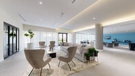Located at 6726 SW 59th Pl in South Miami, this single-floor lounge offers a generously proportioned open-plan layout designed for both relaxed gathering and social interaction. The interior is defined by a refined contemporary aesthetic, with large-format stone-look floor tiles extending throughout the entire space, creating a seamless and cohesive visual flow.

The lounge is organized into several distinct seating zones. A central living area features a substantial sectional sofa paired with sculptural accent chairs arranged atop a patterned area rug, anchored by a wall-mounted TV and a dark display shelving unit. An adjacent zone along the window-lined perimeter offers high-top bistro tables and bar-height seating, bathed in natural light filtered through sheer floor-to-ceiling drapery. A further alcove, distinguished by a bold teal feature wall, provides a more intimate secondary lounge with additional soft seating and a wall-mounted television.

Architectural highlights include a coffered ceiling with integrated warm cove lighting that creates a welcoming ambiance throughout, alongside cylindrical structural columns that add a sculptural quality to the open floor plan. A kitchenette area is visible toward the rear of the space. Potted greenery and curated artwork contribute to the polished, hospitality-inspired atmosphere.