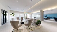 Located at 6726 SW 59th Pl in South Miami, this single-floor lounge offers a generously proportioned open-plan layout designed for both relaxed gathering and social interaction. The interior is defined by a refined contemporary aesthetic, with large-format stone-look floor tiles extending throughout the entire space, creating a seamless and cohesive visual flow.

The lounge is organized into several distinct seating zones. A central living area features a substantial sectional sofa paired with sculptural accent chairs arranged atop a patterned area rug, anchored by a wall-mounted TV and a dark display shelving unit. An adjacent zone along the window-lined perimeter offers high-top bistro tables and bar-height seating, bathed in natural light filtered through sheer floor-to-ceiling drapery. A further alcove, distinguished by a bold teal feature wall, provides a more intimate secondary lounge with additional soft seating and a wall-mounted television.

Architectural highlights include a coffered ceiling with integrated warm cove lighting that creates a welcoming ambiance throughout, alongside cylindrical structural columns that add a sculptural quality to the open floor plan. A kitchenette area is visible toward the rear of the space. Potted greenery and curated artwork contribute to the polished, hospitality-inspired atmosphere.