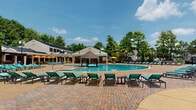 The resort pool area at The Abbys In Inverness in Birmingham, Alabama, offers a thoughtfully designed aquatic and recreational complex that seamlessly blends indoor and outdoor amenities. The centerpiece is a generously sized freeform outdoor pool surrounded by an expansive paver deck, furnished with teal lounge chairs, umbrella-shaded seating, and a centrally positioned gazebo that serves as a striking architectural focal point amid the mature, tree-lined backdrop.

Adjacent to the outdoor pool, a covered indoor pool area provides a sheltered alternative for year-round use. This space features a vaulted ceiling equipped with ceiling fans, open-air column archways that invite natural breezes and views of the surrounding greenery, and a polished aggregate concrete deck furnished with vibrant orange upholstered seating arrangements.

The transition between the indoor and outdoor areas is anchored by a columned breezeway with arched architectural detailing, stone-accented service counters, and raised planter beds filled with colorful seasonal flowers. A welcoming entrance with glass doors and branded signage provides easy access to the facility. Throughout the complex, the combination of open sightlines, lush landscaping, and resort-style furnishings creates a relaxed, upscale outdoor living atmosphere.