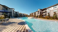 This outdoor amenity space centers around a generously sized, freeform swimming pool with strikingly clear blue water, designed to evoke a resort-style atmosphere. The pool features a gently curving shoreline and includes a central fountain or water feature element, adding visual interest and a sense of luxury to the space. The surrounding deck is finished with an attractive multi-toned paver pattern, combining contrasting shades to create a sophisticated geometric aesthetic underfoot.

The pool area is thoughtfully appointed with blue sling-style lounge chairs and shaded seating arrangements, offering residents multiple spots to relax and enjoy the outdoors. Wrought iron perimeter fencing provides both security and visual definition, while sections of natural wood privacy fencing add warmth and separation between areas. Ornamental grasses and manicured landscaping soften the hardscape and contribute a natural, inviting character to the environment.

The surrounding multi-story residential buildings feature a blend of stone veneer and neutral-toned siding, creating an attractive architectural backdrop. A covered community pavilion or leasing structure with a ceiling fan is visible nearby, reinforcing the functional and social nature of the space. Under clear blue skies, the entire outdoor area benefits from abundant natural light, making it a bright and welcoming communal retreat.