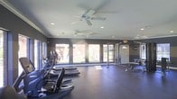 This fitness center is designed to support a comprehensive workout experience within a single-floor layout. The space is anchored by a row of cardio machines, including ellipticals and stair climbers from Vision Fitness, positioned along a wall of large windows that flood the room with natural light and offer views of the surrounding outdoor greenery. The generous window line creates an open, airy atmosphere that makes the space feel expansive and inviting.

The flooring throughout is a durable, dark rubberized material well-suited for high-activity use. Overhead, two ceiling fans are mounted to the smooth white ceiling alongside recessed lighting, ensuring consistent airflow and illumination throughout the room. The walls are finished in a deep charcoal tone, lending the space a sleek, modern aesthetic.

On the opposite side of the room, a strength training area is equipped with a multi-functional cable and rack system, a weight bench, and a dumbbell rack, providing users with a variety of resistance training options. A full-length mirror is positioned along one wall to assist with form and technique. The overall layout is thoughtfully organized, balancing cardio and strength zones while maintaining ample open floor space for movement and stretching.