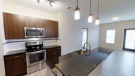 This single-floor residence features three bedrooms and two bathrooms thoughtfully arranged within a well-proportioned floor plan. The heart of the home is a contemporary open-concept kitchen that flows seamlessly into the living area, creating an inviting and functional space for both everyday living and entertaining. The kitchen is outfitted with rich dark-toned cabinetry, sleek granite countertops, and a central island with an undermount sink and a brushed gold faucet. Stainless steel appliances, including a range and over-the-range microwave, add a modern, professional touch. A trio of pendant lights with gold accents hang above the island, providing both task lighting and a warm visual focal point. A track lighting system further illuminates the space from above. The neutral white walls and light flooring keep the aesthetic clean and bright, complementing the darker cabinetry and countertops. A hallway visible from the kitchen leads to the private bedroom spaces, ensuring a clear and practical separation between the living and sleeping areas. The overall design balances modern style with comfortable livability throughout.