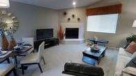This two-story space showcases a contemporary aesthetic with clean lines and a carefully curated design. The living area serves as a focal point, anchored by a sleek, white corner fireplace that adds both warmth and architectural interest. Above the fireplace, a cluster of starburst wall sculptures in metallic finishes adds a bold decorative statement against the neutral gray walls.

The living room is finished with plush wall-to-wall carpeting in a light neutral tone, contributing to a comfortable and cohesive atmosphere. A flat-screen television is mounted on a low-profile black media console, positioned opposite the seating area for optimal viewing. The furniture selection includes a dark leather sofa and modern accent chairs in white upholstery with dark wood frames, complementing the room's monochromatic palette accented with pops of orange.

Natural light filters into the space through a sizable window fitted with horizontal blinds, offering brightness while maintaining privacy. A sleek black coffee table and coordinating side table complete the living area's polished look. The overall ambiance is modern and sophisticated, with thoughtful attention to detail evident throughout the space.