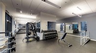 This single-floor fitness center offers a thoughtfully designed workout environment suited for a wide range of exercise routines. The space features a clean, contemporary aesthetic with a neutral color palette of whites and grays, complemented by a durable speckled rubber flooring that provides both safety and comfort underfoot.

The strength training area is well-appointed, housing a full dumbbell rack with a broad selection of weights, a multi-functional cable and weight machine, and an adjustable workout bench, giving users the tools needed for comprehensive free-weight and resistance training. A central structural column subtly divides the space, with large floor-to-ceiling mirrors lining the walls to enhance the sense of openness and allow users to monitor their form.

On the opposite end of the space, a dedicated cardio zone features a row of treadmills and additional aerobic equipment, providing ample options for cardiovascular training. Overhead, linear LED lighting runs the length of the ceiling, ensuring bright and even illumination throughout. The drop-panel ceiling maintains a clean, professional look consistent with a modern fitness facility. Overall, the space is organized, functional, and designed to support a well-rounded fitness experience.