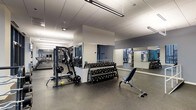 This single-floor fitness center offers a thoughtfully designed workout environment suited for a wide range of exercise routines. The space features a clean, contemporary aesthetic with a neutral color palette of whites and grays, complemented by a durable speckled rubber flooring that provides both safety and comfort underfoot.

The strength training area is well-appointed, housing a full dumbbell rack with a broad selection of weights, a multi-functional cable and weight machine, and an adjustable workout bench, giving users the tools needed for comprehensive free-weight and resistance training. A central structural column subtly divides the space, with large floor-to-ceiling mirrors lining the walls to enhance the sense of openness and allow users to monitor their form.

On the opposite end of the space, a dedicated cardio zone features a row of treadmills and additional aerobic equipment, providing ample options for cardiovascular training. Overhead, linear LED lighting runs the length of the ceiling, ensuring bright and even illumination throughout. The drop-panel ceiling maintains a clean, professional look consistent with a modern fitness facility. Overall, the space is organized, functional, and designed to support a well-rounded fitness experience.