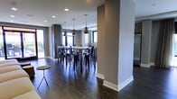 This contemporary single-floor space showcases a thoughtfully designed open-plan layout that blends modern aesthetics with functional living. The interior is anchored by rich, dark hardwood flooring that runs throughout, creating a sleek and cohesive foundation for the space.

A central architectural column adds structural character while naturally defining zones within the open layout. The dining area serves as a focal point, featuring a high communal table surrounded by classic black metal bar-height chairs. Above, a curated arrangement of mixed pendant lights — including industrial cage and dome styles — casts warm, ambient lighting that enhances the modern atmosphere.

The space is bathed in natural light courtesy of large windows on multiple walls, offering views to the exterior and creating an airy, welcoming environment. The neutral gray wall palette complements the dark floors, while crisp white trim and baseboards provide sharp, clean contrast throughout.

A glimpse of an adjacent kitchen area hints at additional functional space, while an access point to an outdoor area is visible, further extending the sense of openness. The overall ambiance is modern, urban, and polished — well-suited for both everyday use and social gatherings.