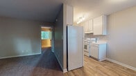 This single-story unit offers a straightforward, functional layout designed for comfortable everyday living. The entry area opens into a living space that features a combination of wood-look hard-surface flooring and plush carpeting, with multiple windows allowing natural light to filter in from the surrounding greenery outside.

The kitchen is efficiently arranged with white cabinetry providing ample upper and lower storage, light-toned countertops, a double-basin sink, and white appliances including an electric range and a full-size refrigerator. A track light fixture overhead ensures the workspace remains well-lit. The kitchen is separated from the main living area by a partial wall, lending a degree of definition between the two spaces.

A hallway connects the living area to the bedroom, which is a generously proportioned room finished with plush carpet flooring and neutral-toned walls. Multiple electrical outlets are positioned throughout the room, offering practical utility.

The bathroom features tile-look flooring, a white tub and shower combination with a chrome showerhead, a toilet, and a vanity with a light-toned countertop. Clean white walls and a small window contribute to a bright, functional atmosphere throughout.