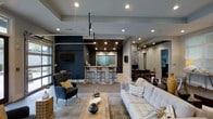 This single-story leasing office showcases a contemporary open-concept design that seamlessly blends functional and welcoming elements. The main lounge area features a light-toned sofa accented with patterned throw pillows, a rustic wood coffee table, and a tufted navy accent chair, creating an inviting reception environment for visitors.

A bold navy blue feature wall anchors one side of the space and supports a wall-mounted flat-screen television, adding a residential feel to the office setting. Adjacent to this lounge area is a bar-height counter with wood-toned stools, illuminated by recessed lighting set into a warm wood-paneled ceiling detail above — providing a distinct and stylish focal point.

The flooring transitions between wood-look planks and carpet, subtly defining different functional zones throughout the space. Recessed lighting is installed throughout the ceiling, ensuring the entire area is well-lit and visually open. A glass-paneled garage-style door on one side allows natural light to filter in, enhancing the airy ambiance.

Toward the rear of the space, additional workstations and seating areas are visible, offering dedicated areas for leasing consultations and administrative tasks. Overall, the office presents a polished, modern aesthetic.