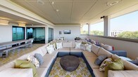 Located at 627 N Lancaster Ave in Dallas, TX, this elevated rooftop lounge offers a sophisticated and comfortable gathering space with sweeping panoramic views of the Dallas skyline and lush treetop canopy. The space is enclosed by a continuous band of large windows that flood the interior with abundant natural light while framing breathtaking views in every direction.

The lounge is thoughtfully divided into distinct functional zones. A generous seating area anchors the main space, furnished with plush wicker sectional sofas and accent chairs arranged around a central coffee table, all anchored by a bold geometric-patterned area rug. This area is ideal for relaxed conversation or social gatherings, and a wall-mounted television adds an entertainment element to the setting.

A long dining area extends along one wing of the space, featuring an extended communal table with bench seating — well-suited for group dining or events. The clean, neutral color palette of cream, gray, and warm wood tones throughout creates a cohesive and inviting atmosphere.

The flooring consists of large-format tile laid in a classic grid pattern, lending a polished and durable foundation to the space. Recessed lighting and built-in audio speakers complement the natural light. The overall ambiance is open, airy, and refined — an exceptional venue for events or everyday use above the Dallas cityscape.