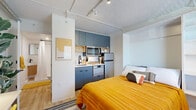 Located at 1104 Avondale Circle in Nashville, TN, this efficiently designed studio offers a smart, modern layout that makes excellent use of its compact footprint. The open main area seamlessly integrates the sleeping, living, and cooking zones into a cohesive, functional space. A built-in Murphy bed with integrated shelving serves as the room's centerpiece, folding away to free up floor space when needed. Light wood flooring and crisp white walls run throughout, creating a bright, airy ambiance complemented by track lighting along exposed ceiling conduits, adding an urban, industrial touch.

The kitchenette is thoughtfully outfitted with slate-blue cabinetry, a full-sized refrigerator, microwave, dishwasher, and a subway tile backsplash. A dedicated workspace with wall-mounted TV provides a practical work-from-home setup. A window allows natural light to filter in, enhancing the open feel of the room.

The bathroom features a clean, modern aesthetic with a walk-in shower, a wall-mounted vanity with warm wood paneling, matte black fixtures, and a corner shelving unit for added storage. The consistent warm gold and white color palette ties the entire space together, delivering a stylish yet highly functional living environment.