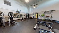 Located at 2302 High Ridge Trail in Fitchburg, Wisconsin, this fitness center is a thoughtfully designed, single-floor facility built to support a comprehensive workout experience. The space features soaring ceilings that lend an open, airy feel, with clerestory windows positioned high on the walls allowing natural light to filter in and brighten the interior. The flooring throughout is durable rubber gym matting, providing both safety and comfort underfoot.

The facility is organized into distinct workout zones. The cardio area houses a well-curated selection of machines, including treadmills, ellipticals, and stationary bikes, all arranged in front of wall-mounted mirrors and flat-screen televisions. The strength training section is equipped with Matrix adjustable benches, a multi-function cable machine, a dumbbell rack with a range of free weights, and various functional fitness accessories such as stability balls, resistance straps, and exercise mats.

The walls combine crisp white paint with an accent of warm terracotta orange and a striking textured brick feature wall, giving the space a modern yet grounded aesthetic. Exposed ductwork adds an industrial character to the upper portion of the room. A glass-panel door leads to an adjoining hallway, ensuring smooth access to the rest of the building. Overall, this fitness center offers a well-rounded, professional-grade environment suitable for a variety of training needs.