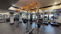 This fitness center is a comprehensively outfitted facility designed to support a full spectrum of training modalities. The space features a striking orange-framed power rack system at its core, complete with a pull-up bar, barbell stations, and a hanging heavy punching bag — ideal for strength and functional training. Surrounding this centerpiece is an extensive selection of selectorized cable machines, a multi-station functional trainer, and adjustable benches positioned throughout the floor. A dedicated dumbbell rack displays a full range of free weights, catering to users of all fitness levels. The cardio zone is equipped with multiple treadmills and upright cycling bikes, each positioned near windows that allow natural light to filter into the space. Wall-mounted flat-screen televisions are integrated along one wall, enhancing the cardio experience. Large mirrors line several walls, amplifying the sense of space and allowing users to monitor their form. The flooring throughout is durable rubber in a speckled pattern, providing safety and comfort underfoot. Recessed lighting and ceiling fans contribute to a well-lit and ventilated environment. The overall design is modern and functional, with a neutral color palette of warm beige walls accented by dark trim and structural columns.