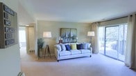 This single-floor space centers around a well-appointed living room that immediately conveys a sense of openness and comfort. The room features soft, neutral-toned walls that complement the plush wall-to-wall carpeting underfoot, creating a warm and cohesive atmosphere throughout. A large sliding glass door fitted with sheer curtain panels floods the space with natural light while providing direct access to an outdoor balcony, where glimpses of lush greenery are visible beyond. The clean white ceiling enhances the bright, airy feel of the room. A structured, light-toned sofa serves as the focal point of the seating area, accented by an array of blue, green, and patterned throw pillows that introduce a pop of color. Flanking the sofa are two tripod-style floor lamps that provide warm ambient lighting. A nautical-themed sailboat painting anchors the main wall, contributing to the space's relaxed, coastal-inspired character. A doorway visible on the left suggests additional rooms beyond, indicating a functional and connected floor plan. Overall, the space balances comfort and style in a clean, livable environment.