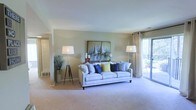 This single-floor space centers around a well-appointed living room that immediately conveys a sense of openness and comfort. The room features soft, neutral-toned walls that complement the plush wall-to-wall carpeting underfoot, creating a warm and cohesive atmosphere throughout. A large sliding glass door fitted with sheer curtain panels floods the space with natural light while providing direct access to an outdoor balcony, where glimpses of lush greenery are visible beyond. The clean white ceiling enhances the bright, airy feel of the room. A structured, light-toned sofa serves as the focal point of the seating area, accented by an array of blue, green, and patterned throw pillows that introduce a pop of color. Flanking the sofa are two tripod-style floor lamps that provide warm ambient lighting. A nautical-themed sailboat painting anchors the main wall, contributing to the space's relaxed, coastal-inspired character. A doorway visible on the left suggests additional rooms beyond, indicating a functional and connected floor plan. Overall, the space balances comfort and style in a clean, livable environment.
