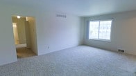 This single-floor space presents a clean, minimalist aesthetic defined by neutral white walls and soft, plush wall-to-wall carpeting in a warm beige tone. The main living area is generously proportioned, creating an open and airy atmosphere that feels welcoming and versatile. A large window allows abundant natural light to fill the room, offering views to the exterior and enhancing the sense of openness throughout the space. The ceiling is smooth and well-maintained, featuring a centrally placed HVAC vent that speaks to a functional, integrated climate control system. A doorway off to one side connects to an adjacent area, suggesting a practical and flowing floor plan. The overall ambiance is bright and understated, providing a neutral canvas well-suited for a variety of interior arrangements. The simplicity of the finishes — from the crisp painted walls to the clean baseboards — gives the space a fresh, move-in-ready character. Whether used as a living area or flexible multipurpose room, the layout and natural light make it an inviting and functional environment.