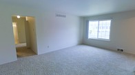 This single-floor space presents a clean, minimalist aesthetic defined by neutral white walls and soft, plush wall-to-wall carpeting in a warm beige tone. The main living area is generously proportioned, creating an open and airy atmosphere that feels welcoming and versatile. A large window allows abundant natural light to fill the room, offering views to the exterior and enhancing the sense of openness throughout the space. The ceiling is smooth and well-maintained, featuring a centrally placed HVAC vent that speaks to a functional, integrated climate control system. A doorway off to one side connects to an adjacent area, suggesting a practical and flowing floor plan. The overall ambiance is bright and understated, providing a neutral canvas well-suited for a variety of interior arrangements. The simplicity of the finishes — from the crisp painted walls to the clean baseboards — gives the space a fresh, move-in-ready character. Whether used as a living area or flexible multipurpose room, the layout and natural light make it an inviting and functional environment.