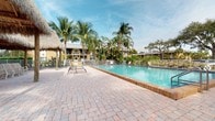 This single-story property is set within a tropical community environment that evokes a distinct Florida resort atmosphere. The standout feature visible from the exterior is a generously sized community swimming pool with clear, blue-green water, framed by a well-maintained brick paver deck that provides ample space for lounging. Cushioned sun chairs are positioned poolside, ideal for relaxing in the abundant natural sunlight. A thatched-roof tiki structure adds a leisurely, vacation-inspired aesthetic to the pool area, offering a shaded retreat. The pool is enclosed by a metal safety fence, and the surrounding landscape is densely planted with tall, swaying palm trees and mature tropical foliage, creating a lush, private feel. The neighboring building facades are finished in warm yellow tones, consistent with the sunny, coastal architectural style typical of the region. The overall ambiance of the outdoor space is relaxed and resort-like, with tropical greenery, open sky views, and well-maintained common areas contributing to a welcoming and leisure-focused environment.