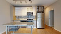 This one-bedroom, one-bathroom residence in Detroit, MI (48226) offers a thoughtfully designed single-floor layout with contemporary finishes throughout. The open-concept kitchen is a standout feature, boasting sleek white cabinetry with glass-front accents, a mosaic tile backsplash, granite countertops, and a full suite of stainless steel appliances including a range, microwave, and refrigerator. Track lighting illuminates the space, highlighting the light wood-tone flooring that flows seamlessly through the main living area. A granite-topped island with a metal frame serves as both a dining and workspace centerpiece, anchoring the transition between the kitchen and the spacious living area. Large sliding glass doors flood the living space with natural light and open to a private balcony, where sweeping views of the surrounding Detroit skyline and a vibrant courtyard below can be enjoyed. The bathroom features a geometric patterned tile floor, a vanity with granite countertops, a vessel sink, and a separate shower area, all finished in a cool, neutral palette. The overall ambiance is modern and urban, combining clean lines, quality materials, and city-connected living in the heart of Detroit.