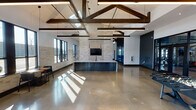 This expansive single-floor amenity space showcases a striking blend of industrial and contemporary design elements. The architecture is defined by soaring vaulted ceilings adorned with prominent exposed dark wood beams, complemented by sleek linear pendant lighting that runs throughout. Polished concrete flooring extends across the entire space, reflecting abundant natural light streaming in through large black-framed windows.

The main gathering area features a prominent stone-clad fireplace wall with a mounted flat-screen television, flanked by a sophisticated dark navy reception or service counter with a light stone top. A lounge zone is furnished with modern seating, a distinctive gold accent coffee table, and an area rug, creating a comfortable social hub. Stone tile wainscoting runs along the lower walls, adding texture and warmth.

A dedicated recreational zone includes a foosball table and additional casual seating arrangements. The space also incorporates a fitness center, accessed through glass-paneled navy-framed double doors, visible with cardio equipment inside. A richly toned dark wood accent wall adds depth near the fitness entrance. Throughout, the navy blue color palette, natural stone accents, and industrial-inspired details create a cohesive, polished atmosphere.