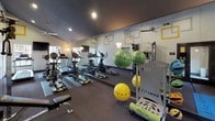 Located at 1500 S Jupiter Rd in Allen, Texas, this single-floor fitness center offers a well-equipped and thoughtfully designed space for a comprehensive workout experience. The interior is distinguished by its modern aesthetic, featuring bold yellow and gray geometric square motifs adorning the white walls, creating a vibrant and energizing atmosphere. Soaring vaulted ceilings with recessed lighting and ceiling fans contribute to the open, airy feel of the space, while large windows allow natural light to filter in and provide views of the surrounding outdoor environment. A full wall of mirrors along one side of the room amplifies the sense of space and allows users to monitor their form during exercise. The cardio area is well-stocked with multiple treadmills, elliptical machines, and stationary bikes, all positioned for easy access. A dedicated strength training zone includes a cable machine, weight bench, resistance accessories, and stability balls stored on a fitness rack. Wall-mounted flat-screen televisions are positioned throughout the room for entertainment during workouts. The flooring transitions between durable rubber matting and carpeted sections, providing appropriate surfaces for both equipment use and floor exercises. A yoga mat and recovery tools are also available, rounding out the facility's offerings. The space is accessible via glass-paneled double doors.