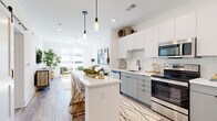 This single-floor residence showcases a thoughtfully designed open-concept layout with clean, modern aesthetics evident in every room. The kitchen serves as a central hub, featuring flat-front cabinetry in white and soft grey tones, a white quartz waterfall island with seating, subway tile backsplash, stainless steel appliances, and sleek black fixtures. Pendant lights hang above the island, adding a refined touch. A sliding barn door accent near the entryway introduces a subtle industrial element alongside the bold blue entry door.

The living area flows seamlessly from the kitchen, bathed in natural light from large windows. Light wood-tone flooring runs throughout, creating visual continuity. The living room accommodates a comfortable seating arrangement beneath a crystal chandelier, with ample room for additional furnishings.

The bedroom is bright and airy, benefiting from generous window views and a ceiling fan. It offers comfortable proportions with light wood flooring and crisp white walls.

The bathroom is finished to a high standard, featuring large-format marble-look tile flooring, a glass-enclosed walk-in shower with full-height tile surround, a white vanity with black fixtures, built-in niche shelving, and an LED-backlit mirror, creating a clean, spa-inspired atmosphere.