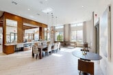 Located at 2926 Barker Cypress Rd in Houston, TX, this single-floor community clubhouse is thoughtfully designed to serve as a welcoming gathering space. The open-plan layout is anchored by soaring ceilings that amplify the sense of spaciousness, while large floor-to-ceiling windows and glass panel doors flood the interior with generous natural light and provide inviting views of the surrounding outdoor areas.

The main gathering area features light-toned, large-format tile flooring that runs throughout, creating a clean and cohesive aesthetic. A substantial dining table with upholstered chairs serves as a central focal point, suspended above by elegant globe pendant lighting that adds a warm, contemporary touch. Adjacent to the dining area, a shuffleboard table offers a recreational amenity, complementing the social atmosphere of the space.

A dedicated wet bar and kitchenette zone, framed by rich warm-wood cabinetry, provides functional amenities for entertaining. Nearby, a well-appointed mailroom features an extensive bank of individual resident mailboxes, underscoring the space's role as a community hub. Smaller bistro-style seating arrangements are positioned near the windows and exterior doors, offering casual spots for conversation. Double glass doors open directly to an outdoor courtyard, seamlessly connecting the interior to the exterior environment.