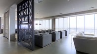 This single-floor lounge space showcases a refined contemporary design that balances openness with thoughtful architectural detailing. One of its most striking features is the floor-to-ceiling glass window wall spanning the perimeter, flooding the interior with abundant natural light and framing expansive panoramic views of the surrounding landscape. Sheer vertical blinds filter the incoming light, creating a soft, ambient glow throughout the space.

A standout architectural element is the bold, dark-toned decorative lattice screen partition, which features intricate geometric patterning. This partition serves both as a visual divider and an artistic focal point, adding depth and texture to the otherwise clean-lined interior. The polished concrete flooring reinforces the modern aesthetic, providing a sleek, neutral foundation that complements the overall palette.

The seating area is furnished with multiple groupings of structured grey sofas and armchairs arranged to encourage conversation and relaxation. Pendant lighting fixtures are suspended from the ceiling at varying intervals, contributing warm accent lighting that enhances the lounge atmosphere. Recessed ceiling lights provide even, functional illumination throughout. The overall ambiance is modern, sophisticated, and inviting.