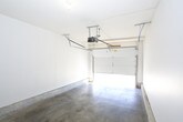 This single-level garage is a straightforward, functional space designed to accommodate vehicle storage and utility needs. The floor throughout is bare concrete, providing a durable and easy-to-maintain surface well-suited for a variety of uses beyond vehicle parking, including storage or workshop activities.

The garage is equipped with a sectional overhead door fitted with an automatic opener and standard rail track system, allowing convenient vehicle access. When open, the door reveals views of the surrounding outdoor area, including a glimpse of landscaping beyond the entrance.

The interior walls are finished in clean white paint, giving the space a bright and open feel despite its utilitarian purpose. Two wood-toned interior doors are positioned along the rear wall — one leading to an adjacent interior space and another providing secondary access. An electrical panel is mounted on the rear wall between the two doors, centralizing utility access within the space.

Overhead, the ceiling is smooth and unfinished, with a basic light fixture providing illumination. The overall layout is open and uncluttered, offering ample floor space for flexible use. The combination of the automatic door system, concrete flooring, electrical panel, and interior access points makes this garage a practical and well-equipped space for everyday functional needs.