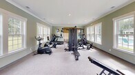 Located at 102 Lusk Lane in Pooler, GA, this well-appointed fitness center offers a comprehensive workout environment across two dedicated exercise rooms. The space is finished with a calming sage-green color palette, white crown molding, and neutral carpeted flooring that lends a clean, inviting atmosphere throughout.

The main exercise room is generously proportioned, accommodating a wide array of equipment including elliptical trainers, stationary bikes, a rowing machine, a treadmill, and a multi-station cable strength machine, along with a weight bench and dumbbell rack. Multiple large white-framed windows line the walls on both sides, flooding the room with natural light and offering pleasant outdoor views of the surrounding greenery and pool area. Recessed ceiling lighting provides additional illumination, and crown molding adds a refined architectural touch.

The secondary exercise room offers a more intimate setting, ideal for stretching, free weights, or focused individual workouts. It features its own window for natural light and direct exterior access through a glass-paneled door.

A built-in bench and cubby area serves as a convenient transitional space between the two rooms, providing a spot to store personal belongings. Framed artwork adorns the walls throughout, enhancing the overall ambiance. This fitness center combines functionality with thoughtful design details to create a comfortable and motivating environment.