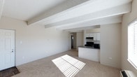 This single-floor space offers a clean, open-plan layout that seamlessly connects the living area to the kitchen. The living space is finished with wall-to-wall carpet that adds warmth underfoot, while the textured ceiling features distinctive exposed structural beams that lend a subtle architectural character to the interior. Natural light streams in through a window, casting bright patterns across the floor and giving the space a fresh, airy feel.

The kitchen is positioned at the far end of the open living area, defined by a peninsula countertop finished in dark laminate — a practical and visually grounding feature that separates the cooking space from the main living zone. White upper cabinetry lines the kitchen walls, providing ample storage while keeping the palette light and cohesive. Black appliances, including a range with overhead ventilation hood and a microwave, provide a contrasting accent. A hallway visible beyond the kitchen suggests additional rooms or utility spaces toward the rear of the floor plan.

The overall color palette is neutral throughout — soft whites, warm beiges, and subtle grays — creating a versatile backdrop well-suited to a variety of interior styles. The combination of open sightlines, natural light, and functional kitchen design makes this a practical and inviting single-level space.