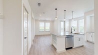 Located at 405 E Ash St in Celina, TX, this thoughtfully designed one-bedroom, one-bathroom residence offers a clean, modern aesthetic across its single-floor layout. The open-concept kitchen and living area serves as the heart of the home, showcasing a large center island topped with granite countertops and illuminated by pendant lighting. White shaker-style cabinetry, a subway tile backsplash, and stainless steel appliances — including a refrigerator, range, and built-in microwave — complete the well-appointed kitchen. Light hardwood-style flooring runs throughout the main living areas, enhancing the bright, airy ambiance created by multiple windows with outdoor views of lush greenery. The bedroom offers plush carpeting, a ceiling fan, and generous natural light through dual windows. Direct access leads to a dedicated closet for convenient storage. The bathroom is a standout feature, presenting a double-sink vanity with granite countertops, black matte hardware, framed mirrors, and a glass-enclosed walk-in shower with elegant tile surround. Light wood-look flooring ties the space together seamlessly. Recessed lighting throughout adds a polished, modern touch to this well-designed Celina residence.