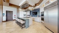 Located along South Ocean Boulevard in Pompano Beach, Florida, this generously proportioned single-floor space offers a sophisticated coastal aesthetic defined by refined architectural details and an abundance of natural light. Throughout, large-format marble tile flooring in warm cream tones creates a seamless, polished foundation, while dramatic soaring ceilings with warm wood accents lend a sense of grandeur.

Floor-to-ceiling windows and glass doors line multiple walls, framing lush tropical palm views and providing direct access to an outdoor terrace. The open layout accommodates several distinct areas: a grand lounge featuring a crystal chandelier, marble-clad columns, a fireplace, and multiple seating arrangements with plush upholstered furniture; a casual sitting area with woven rattan chairs positioned to take full advantage of the sweeping outdoor views; and a formal dining room enclosed by glass doors, appointed with a tray ceiling featuring a distinctive blue inlay, a pendant lantern fixture, and a substantial wood dining table surrounded by patterned upholstered chairs.

The kitchen is a standout feature, showcasing custom white cabinetry, a striking blue mosaic tile backsplash, stainless steel appliances, double ovens, a gas cooktop, and a large island with a dramatic green marble waterfall countertop and bar seating. Built-in shelving and a billiards table round out the versatile amenity offerings of this well-appointed coastal gathering space.