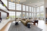 Located at 4720 Nations Crossing Rd in Charlotte, NC, this single-floor living space makes a striking architectural statement through its dramatic double-height, floor-to-ceiling grid windows that flood the interior with abundant natural light and frame sweeping views of the surrounding greenery. The soaring ceilings lend an airy, expansive quality to the space, complemented by warm hardwood flooring and a clean white palette throughout.

The living room serves as the heart of the space, anchored by a bold black-and-white geometric area rug and an eclectic mix of contemporary furnishings, including a channeled olive-green sofa, sculptural accent chairs, and organic coffee tables. Pendant lighting with artistic globe fixtures adds warmth and visual interest overhead.

An entire wall is dedicated to a custom built-in shelving unit with integrated cabinetry and media storage, offering both function and curated display space. A sleek glass-paneled interior door with black framing and brass hardware provides an elegant transition between areas.

A standout wet bar nook features sage green cabinetry, dark stone countertops, wine storage, and a breathtaking iridescent penny tile accent wall illuminated by gold pendant lights — a truly distinctive design feature that elevates the overall aesthetic of the space.