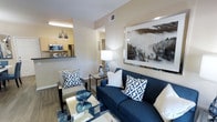 This single-floor residence offers a well-appointed living space that seamlessly blends comfort and modern style. The living area serves as the heart of the home, anchored by a bold navy sofa accented with geometric-patterned throw pillows and complemented by a sleek mirrored coffee table. A large framed landscape artwork depicting a misty forest adds a striking focal point to the neutral-toned walls, enhancing the room's calm and contemporary ambiance. Warm hardwood-style flooring runs throughout, adding continuity and a sense of warmth to the space. An accent chair with matching geometric cushions and a glass side table complete the seating arrangement, creating an inviting and functional area for relaxing or entertaining. The living space flows naturally into the kitchen, which is visible through an open pass-through counter. The kitchen features light wood cabinetry, stainless steel appliances including a microwave and refrigerator, and dark countertops that provide an elegant contrast. Recessed and overhead lighting ensures the entire space is well-lit. Overall, the residence presents a cohesive, modern aesthetic with thoughtful design details throughout.