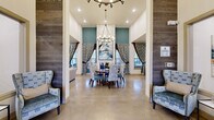 Located at 5803 SW Parc St in Bentonville, Arkansas, this single-story clubhouse is a thoughtfully designed communal facility that blends contemporary style with welcoming functionality. Polished concrete floors run throughout, providing a cohesive foundation that ties together the various distinct zones within the open layout.

The central gathering area features soaring ceilings and an elegant chandelier above a round dining table framed by upholstered chairs, flanked by wood-paneled accent walls and dramatic patterned drapery. Adjacent to this is a spacious conference-style dining room with a tray ceiling finished in deep teal, accommodating a large rectangular table and an abundance of natural light through generously sized windows overlooking the outdoors.

A well-appointed kitchen area serves as a social hub, centered around a substantial island with bar seating, pendant lighting, and stainless steel appliances. Multiple lounge areas offer comfortable seating arrangements with bold accent walls, flat-screen televisions, and curated artwork. A dedicated business center provides computer workstations and printing equipment in a quietly styled nook. The cohesive teal, gold, and neutral color palette lends the entire space a polished, resort-inspired ambiance.