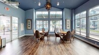 This single-floor community clubhouse is thoughtfully designed to provide residents with an inviting and functional gathering space. The interior is defined by its striking floor-to-ceiling window walls, which flood every area with abundant natural light and offer unobstructed views of the outdoor pool deck, pergola, and the surrounding residential neighborhood. The rich, dark hardwood-style flooring runs throughout the space, providing a warm contrast to the cool blue-grey tones of the walls and ceilings.

The lounge area is generously proportioned, featuring a comfortable seating arrangement with a neutral-toned sofa, accent chairs, glass-top coffee tables with gold accents, and elegant floor lamps — creating a sophisticated yet welcoming atmosphere. A separate dining area showcases soaring vaulted ceilings painted in a bold blue, anchored by a dramatic orb chandelier. Round dining tables surrounded by leather accent chairs offer casual seating, while oversized windows frame views of the outdoor amenity area beyond.

The 3D layout reveals a well-organized floor plan that includes a stone-accented feature wall, a built-in bar or service area, and multiple distinct zones for lounging, dining, and socializing. Wall sconces and pendant lighting provide warm accent illumination throughout, enhancing the overall ambiance of this elegantly appointed community space.