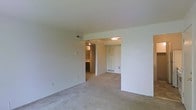 This single-floor residential space offers a straightforward and functional layout designed for comfortable everyday living. The main living area is finished with wall-to-wall neutral carpeting that extends throughout, providing a soft and cohesive feel underfoot. Smooth, white walls create a clean, bright backdrop that allows for easy personalization and décor.

A structural beam subtly defines the transition between the main living space and the adjoining areas, adding architectural interest to the open layout. Visible from the living area, a doorway leads into what appears to be a kitchen or utility space, offering a glimpse of cabinetry and overhead lighting. Nearby, a white interior door provides access to additional rooms or storage, contributing to the overall sense of connectivity within the floor plan.

Practical details such as baseboard heating vents and a wall-mounted thermostat reflect the functional infrastructure of the space. Recessed overhead lighting in the connecting area ensures adequate illumination throughout. The neutral color palette, open sightlines, and straightforward design make this a versatile space well-suited to a variety of living arrangements.